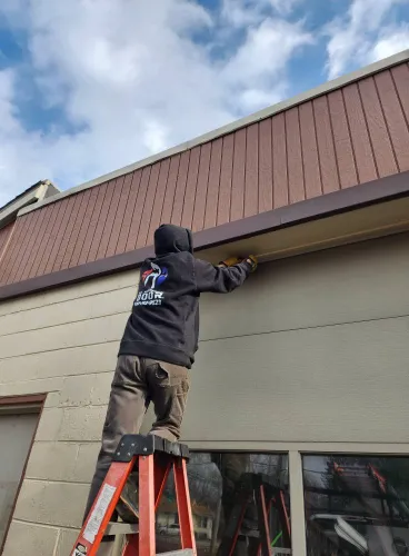 Man wearing a All American Door company sweater stands on top of a ladder performing a garage door repair service.