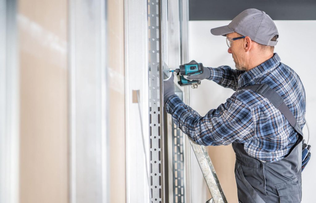 A skilled worker in checkered polo under an overall secures the garage door screws with a screwdriver.