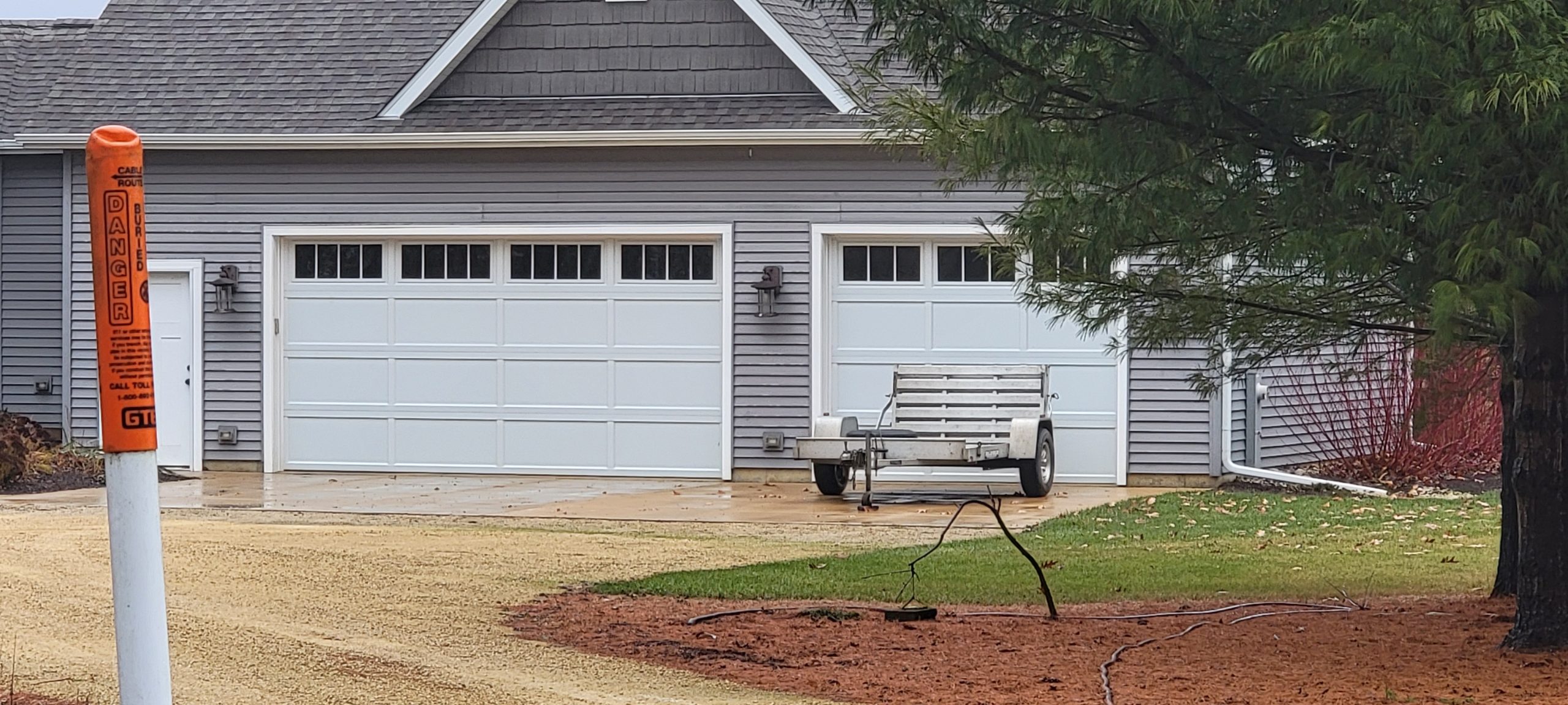 Gray garage with double doors, a caution sign, and a trailer on a gravel driveway
