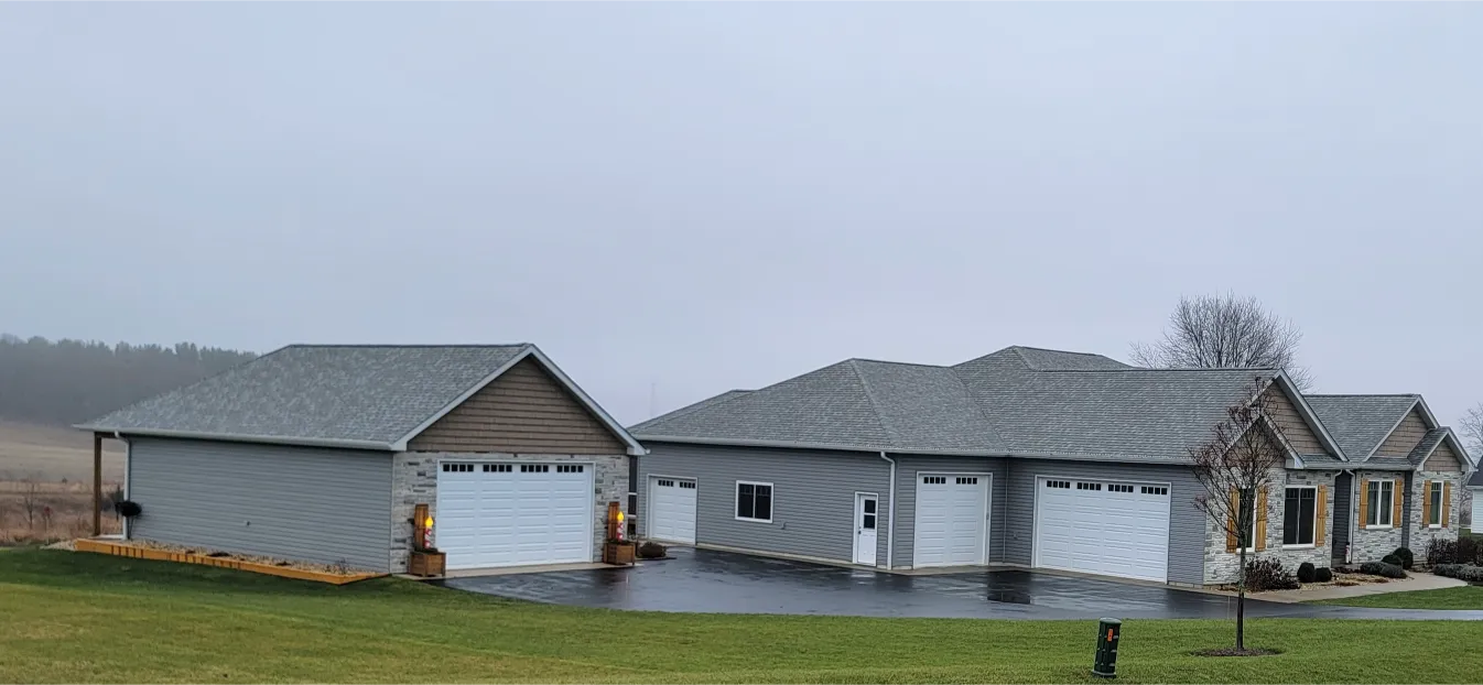 A modern ranch-style house with multiple garages, set on a cloudy day, surrounded by a green lawn and open countryside in the background.