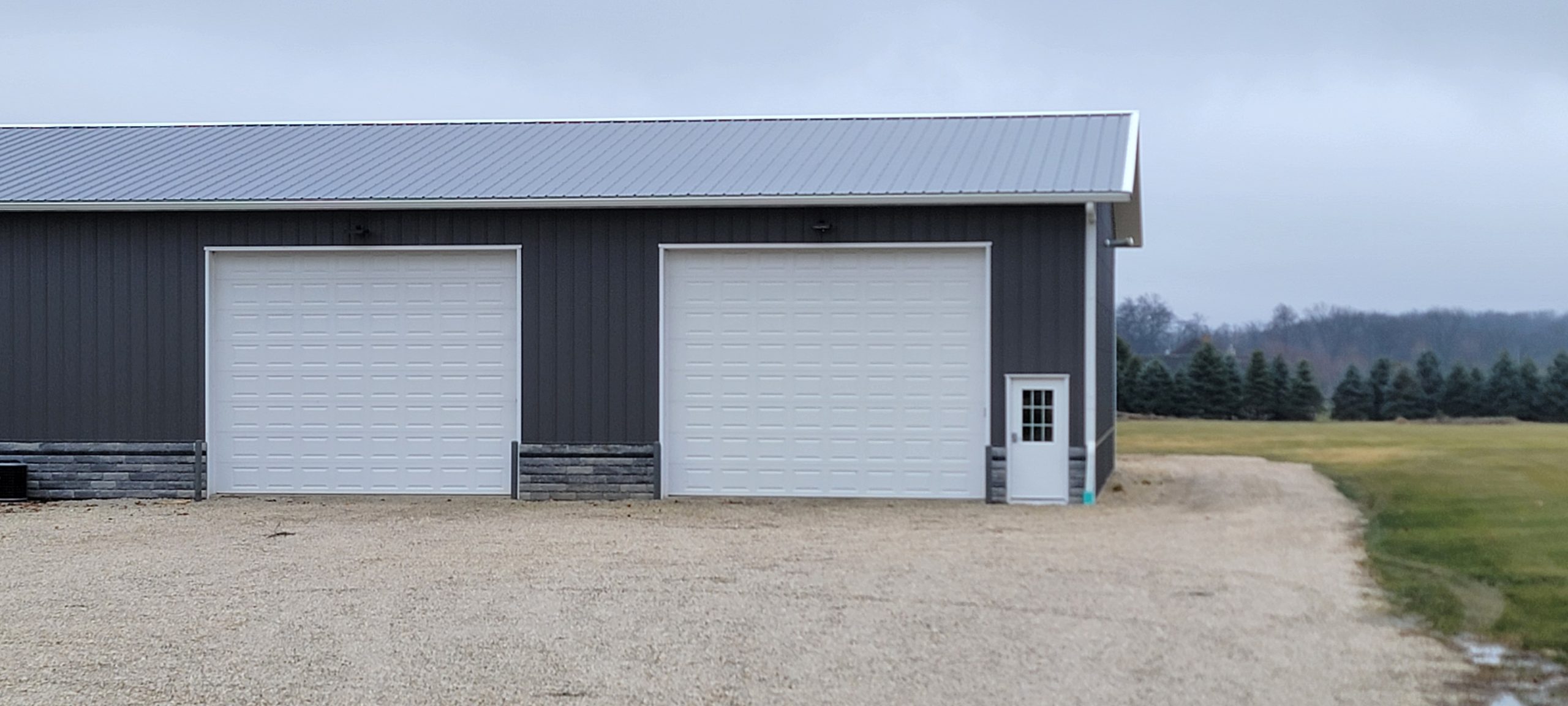Large gray metal garage with two white doors and a small side door, surrounded by a gravel driveway and grass field.
