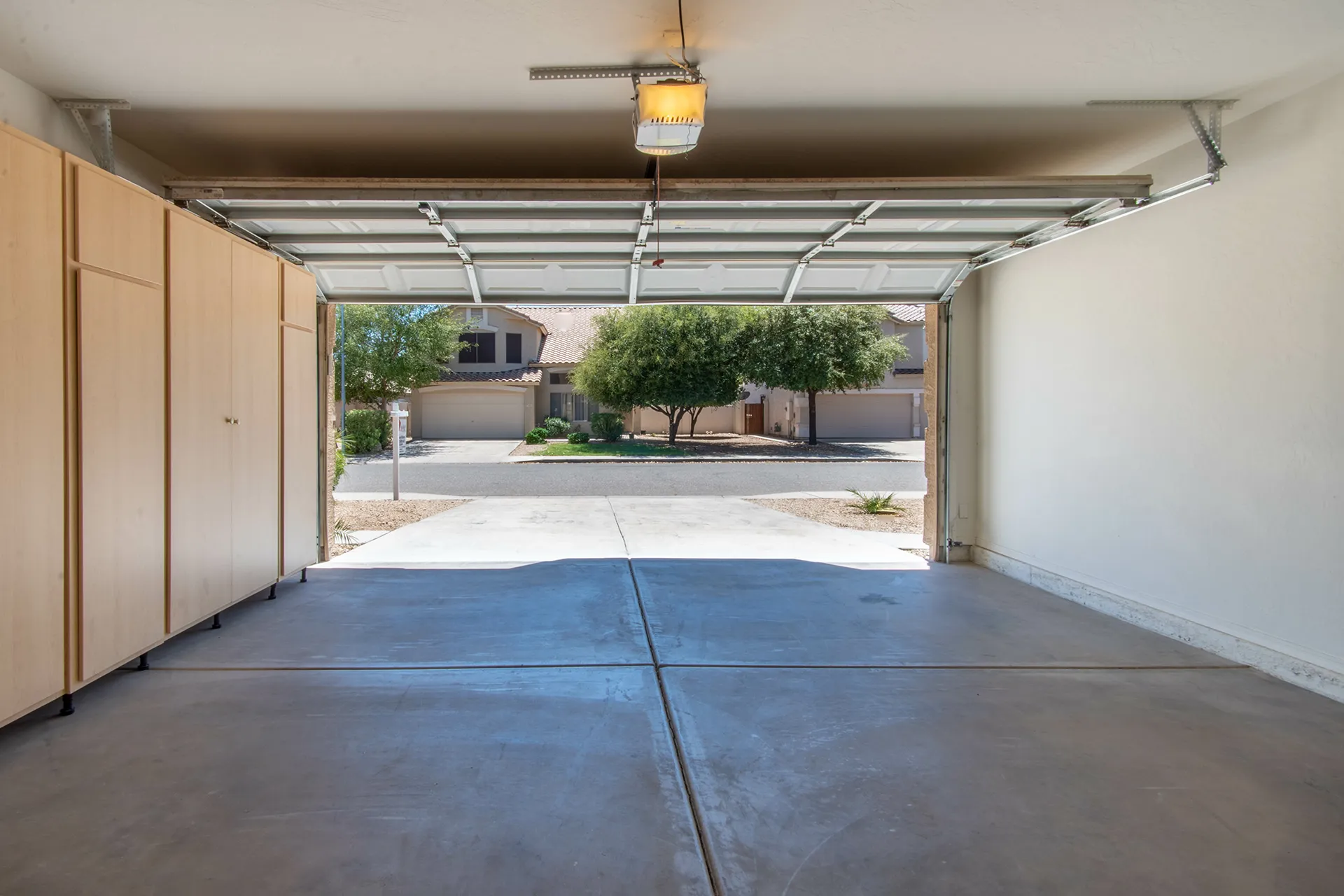 View from inside an open garage with light wooden cabinets on the left, facing a quiet neighborhood street with trees and houses.